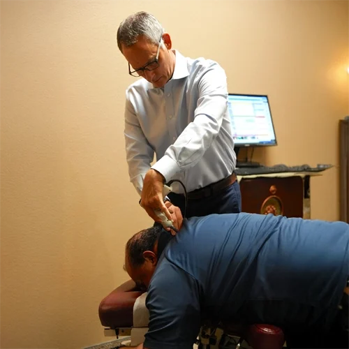 Dr. Michael Ganschow performing a targeted chiropractic adjustment on a patient’s upper back using a handheld device, with a computer workstation visible in the background.
